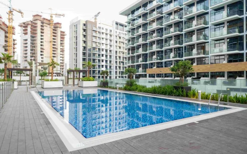 Outdoor swimming pool with clear water, surrounded by modern apartment buildings and greenery, featuring recent Pool Area Upgrades and construction cranes visible in the background.