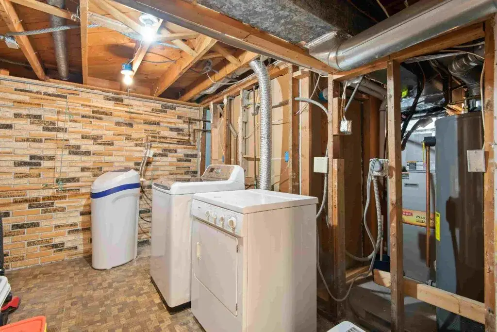 Unfinished basement laundry area with exposed ceiling beams, washing machine, dryer, water softener, visible ductwork and pipes, and Poly B Plumbing throughout.