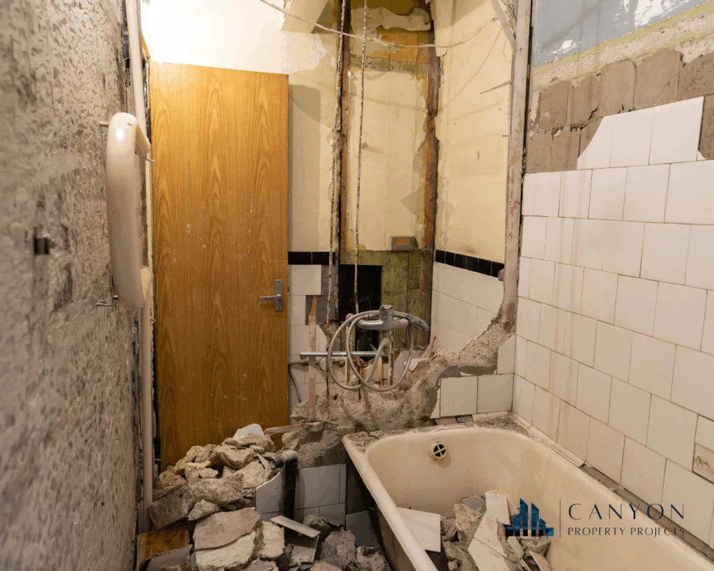 A bathroom with broken tiles, rubble on the floor, exposed pipes, and a damaged bathtub; part of the wall is demolished—ideal for bathroom renovation contractors. Canyon Property Project logo in the corner.