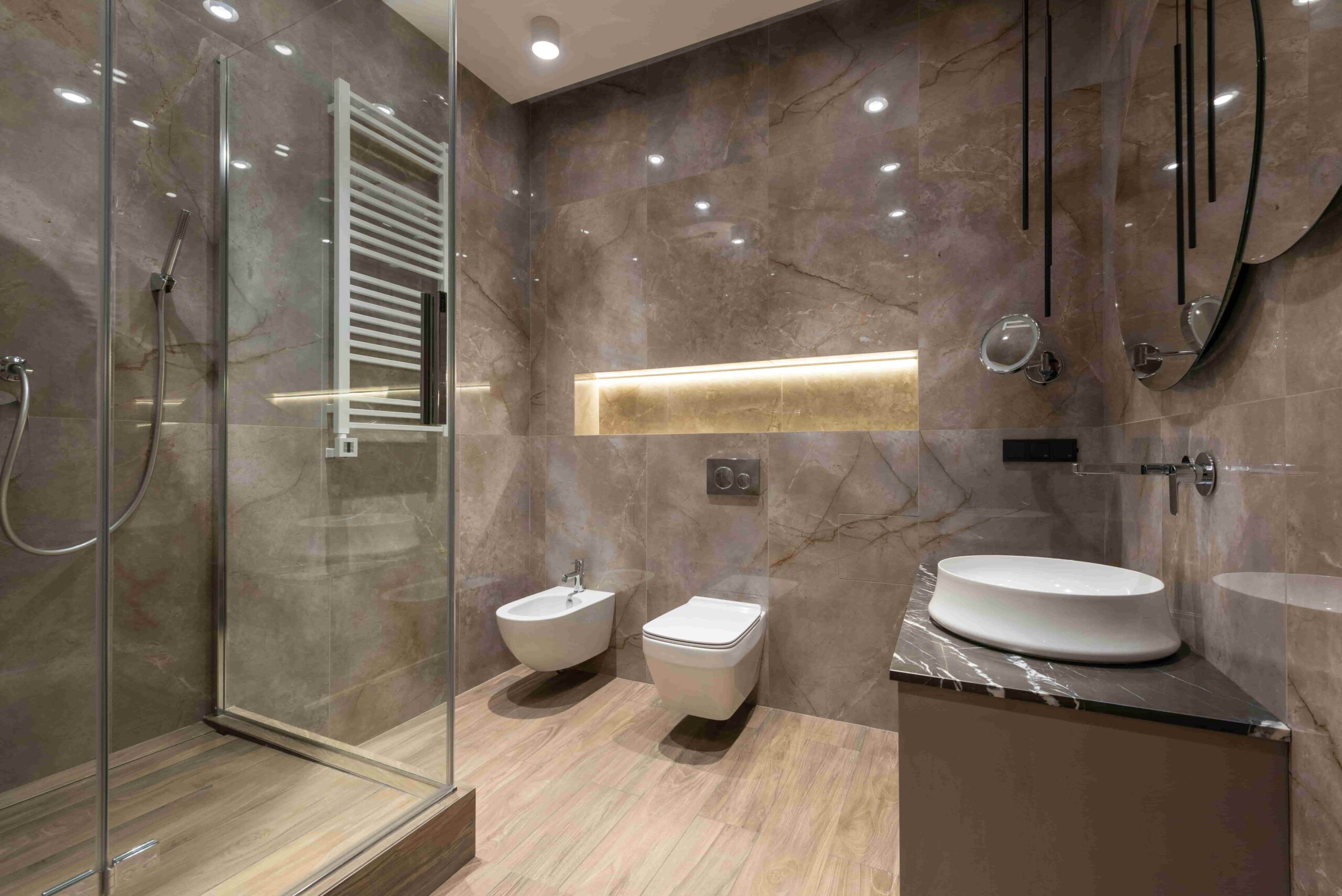 Modern bathroom with marble walls, wood-look floor, glass shower, wall-mounted toilet and bidet, vessel sink on a vanity, large mirrors, and illuminated shelf—perfect inspiration if you’re wondering How Long Does a Bathroom Renovation Take in New Westminster?.