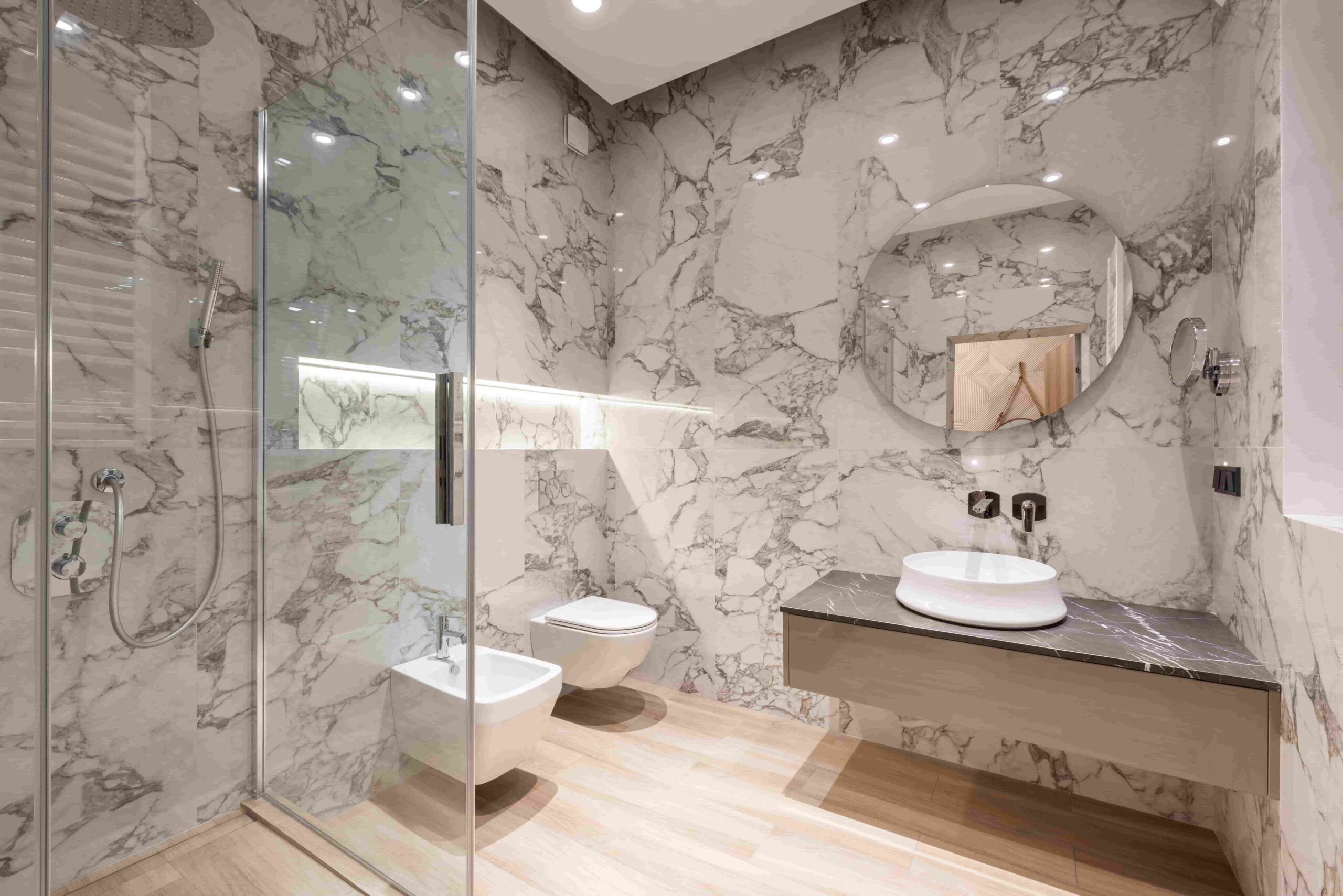 Modern bathroom with marble walls—one of the best materials to use for a bathroom renovation in a Richmond home—plus a glass shower, wall-mounted toilet and bidet, round mirror, floating vanity with vessel sink, and wood-look flooring.