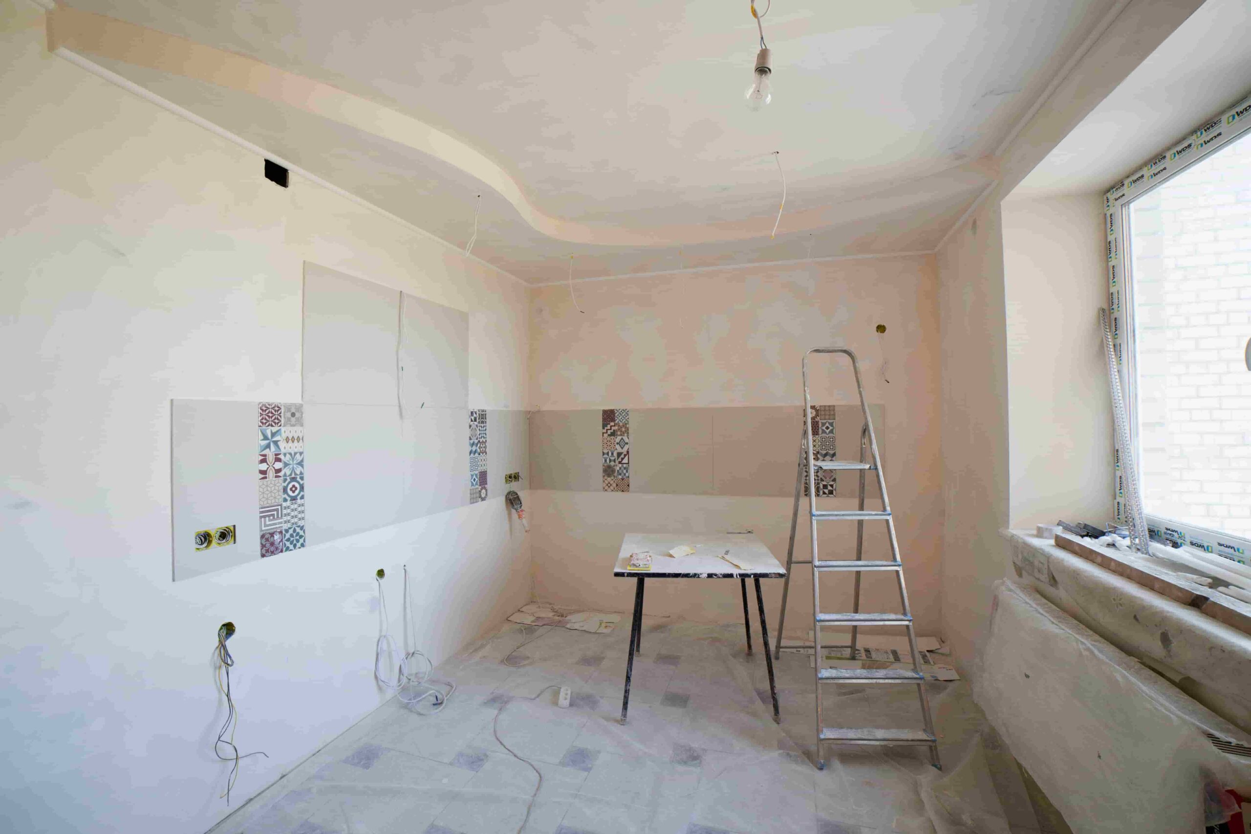 A room under renovation with unfinished walls, a step ladder, a table with tools, exposed wiring, and a large window letting in natural light—perfect inspiration for budget-friendly home renovation ideas for Coquitlam homes.
