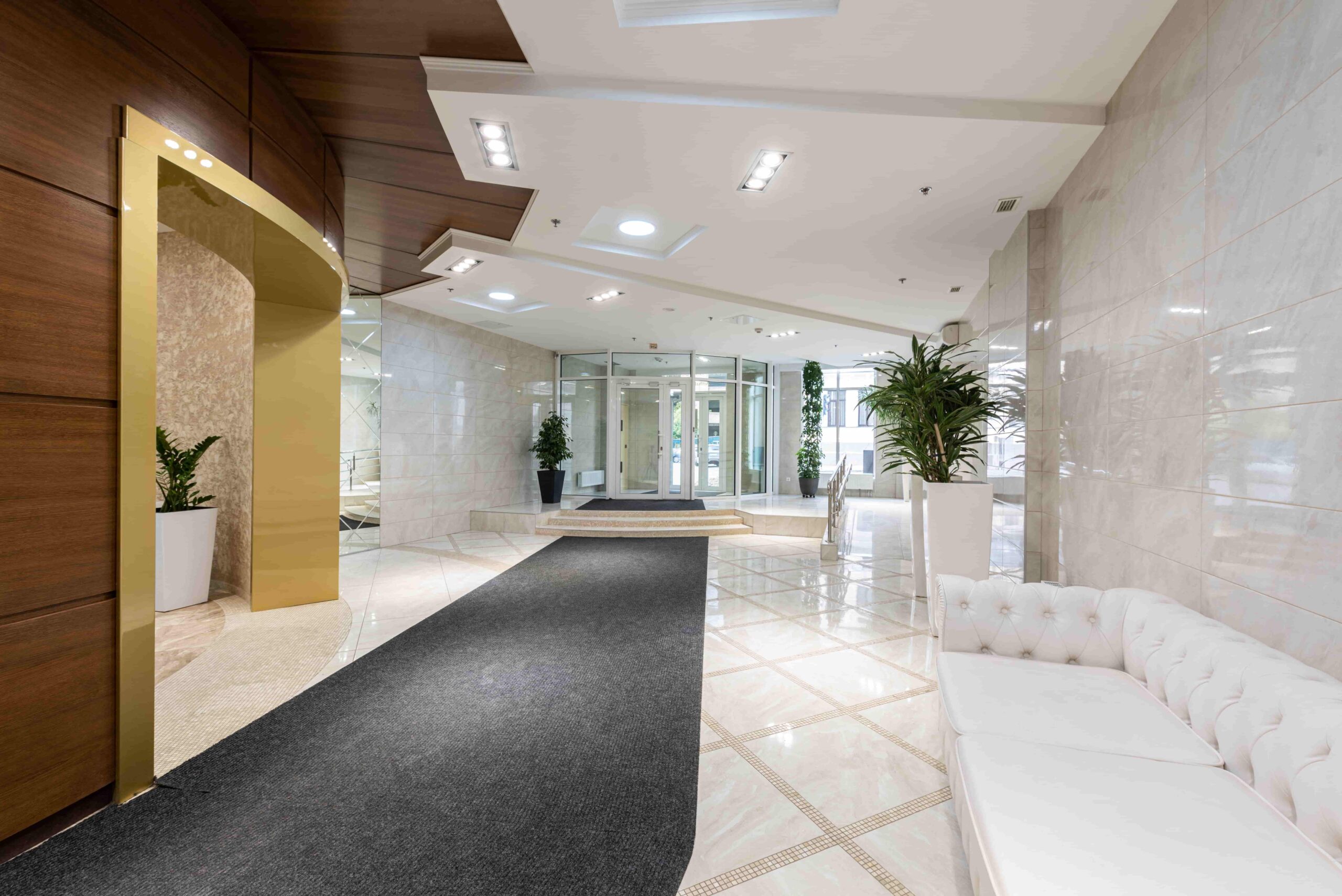 Modern office lobby with marble floors, white sofa, potted plants, and glass entrance doors; wood and gold accents on the walls showcase stunning multifamily lobby transformations.