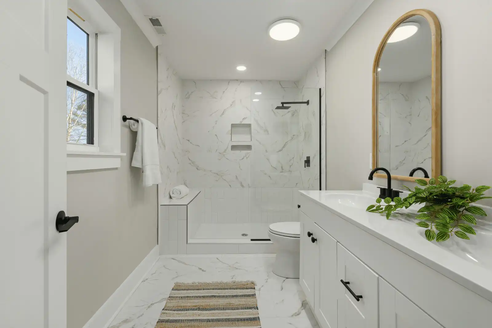 Modern bathroom with white marble shower, glass door, white vanity with black fixtures, oval mirror, potted plant, and striped rug on white tile floor. Towel hangs by the window.