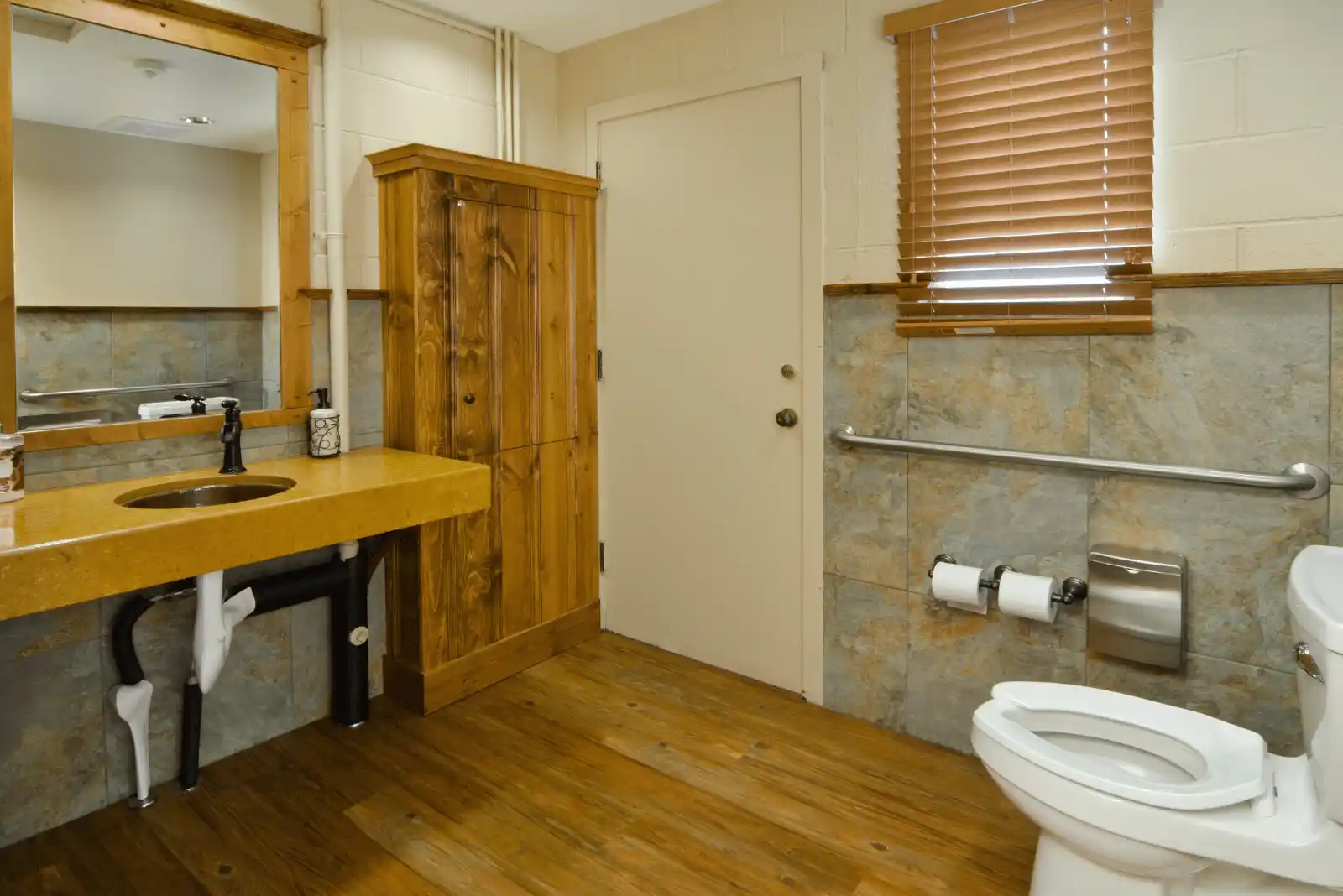 Accessible bathroom with wooden floor, sink with open plumbing, mirror, cabinet, toilet with grab bar, two toilet paper rolls, and window with blinds.