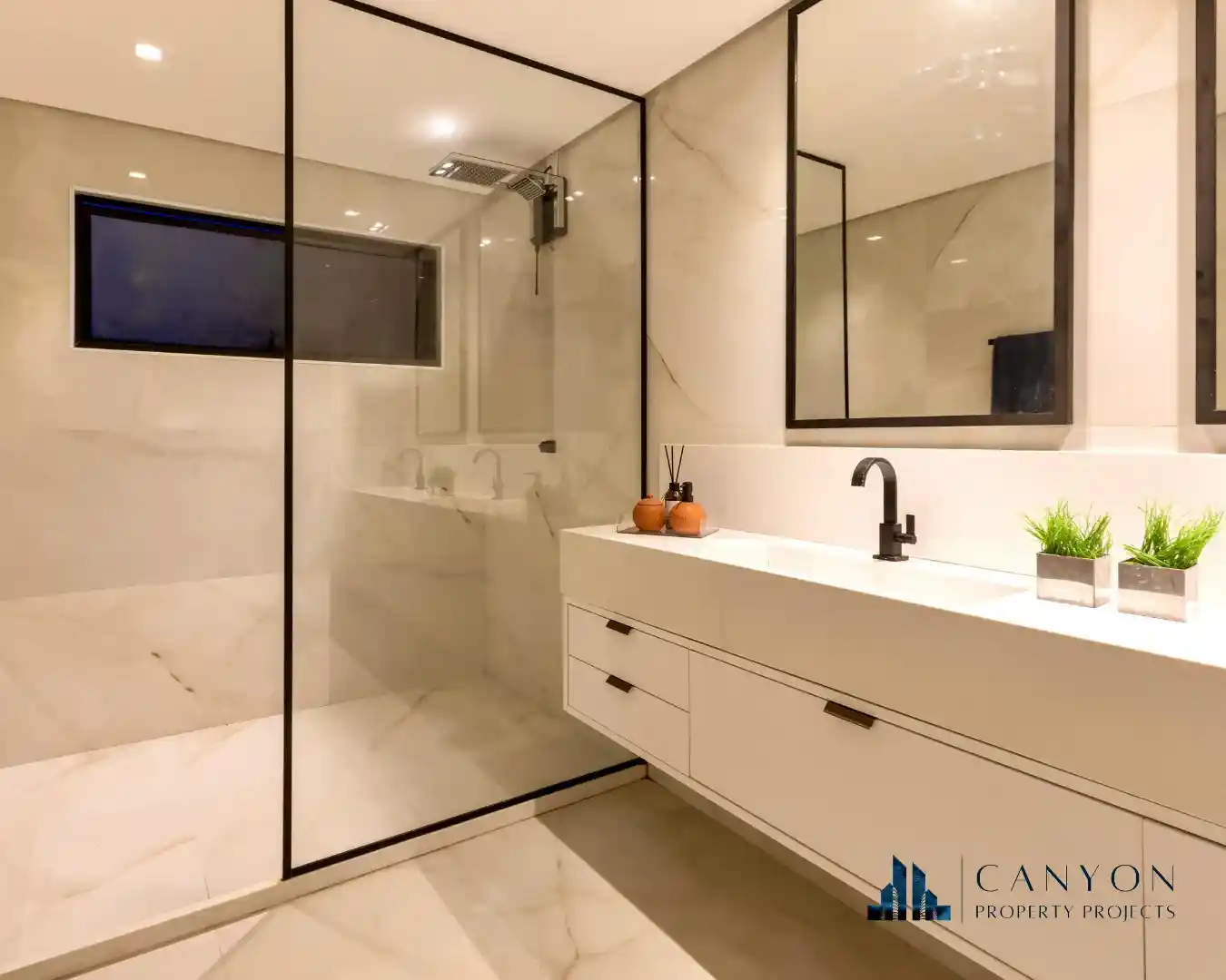 Modern bathroom with a large glass-enclosed shower, white vanity with black fixtures, two mirrors, and potted plants on the counter—showcasing the quality of Strata Property Services. Canyon Property Projects logo in the corner.