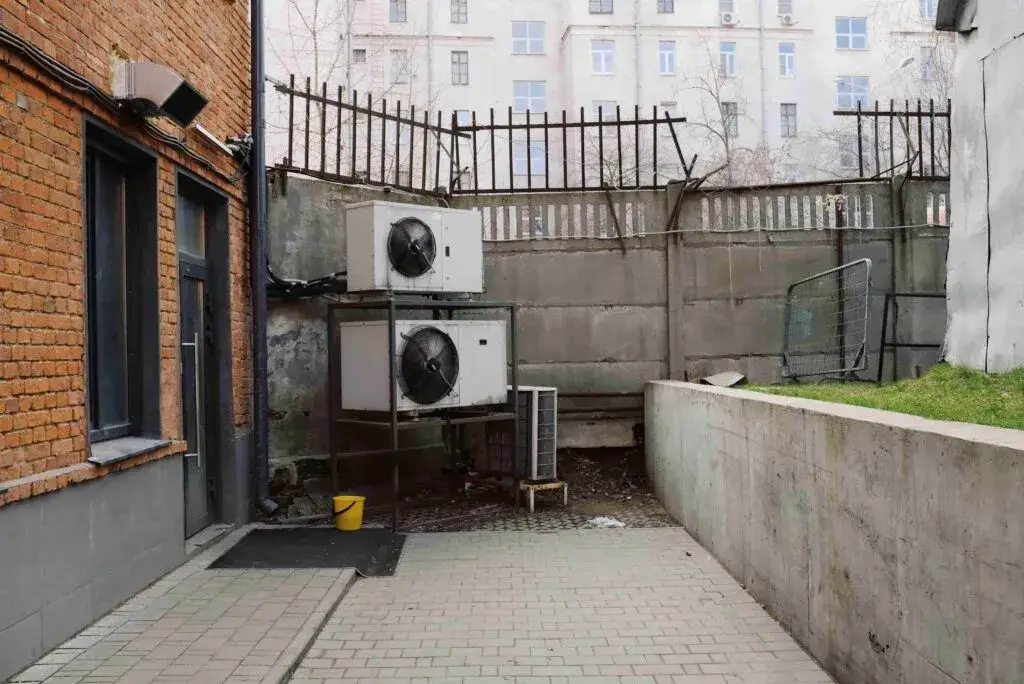 Two large outdoor air conditioning units, part of recent HVAC upgrades, are stacked on a metal frame next to a brick building, with a yellow bucket on the ground nearby and a concrete wall in the background.