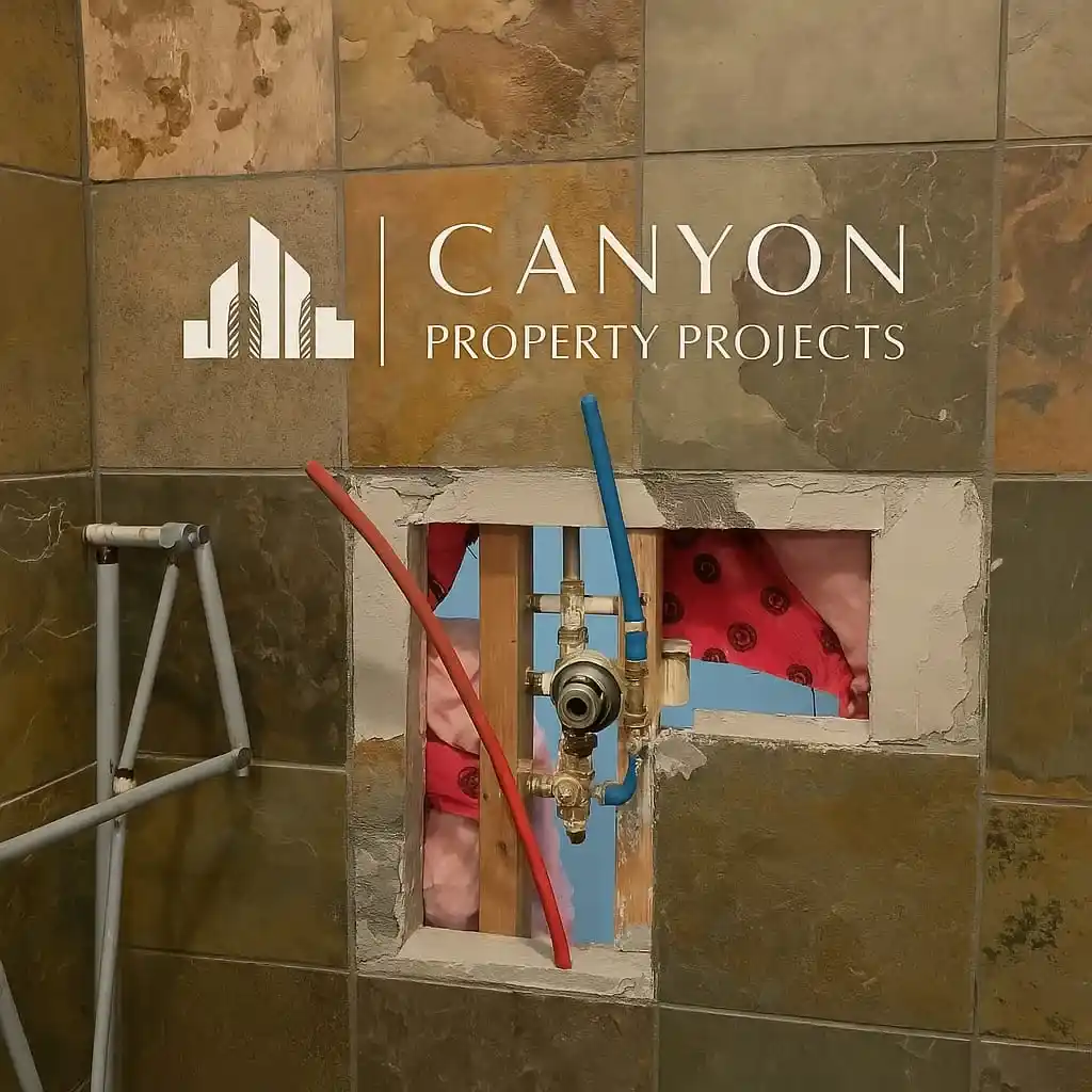 A tiled wall with an exposed plumbing fixture and insulation, showing a square opening and some pipes during a polybutylene pipe replacement; Canyon Property Projects logo is overlaid.
