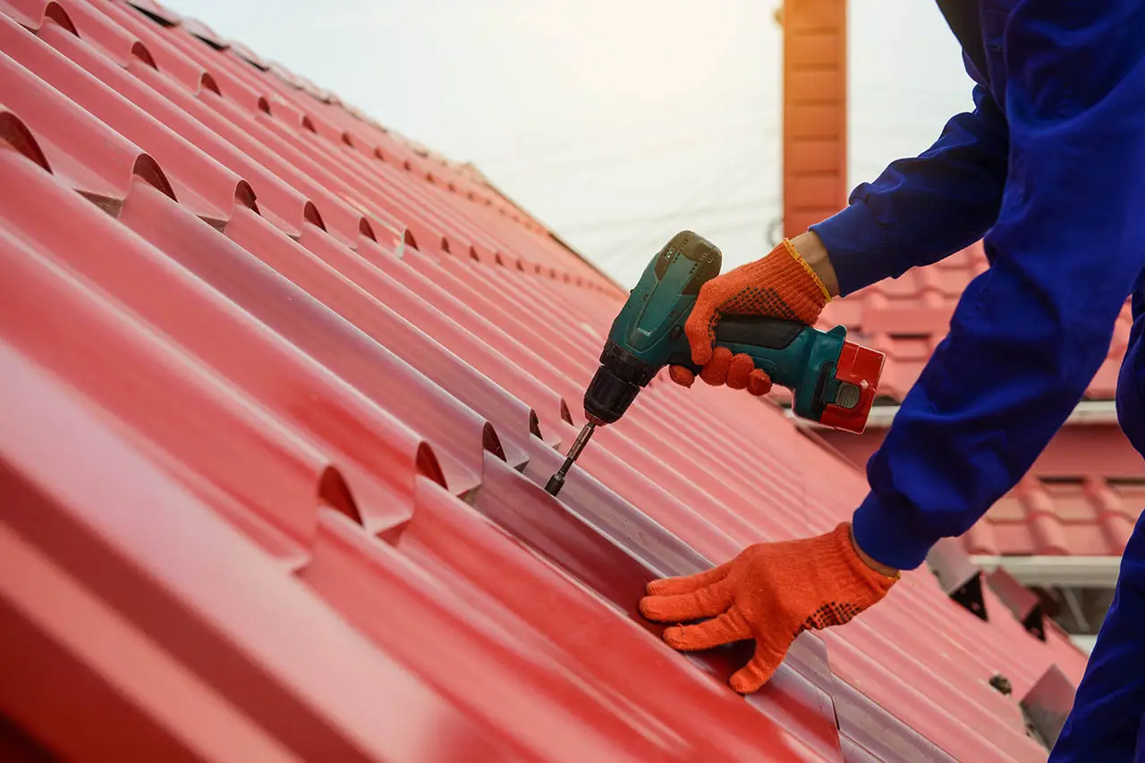Person wearing gloves uses a power drill to fasten a red metal roofing sheet on a sloped roof during a roof replacement project.