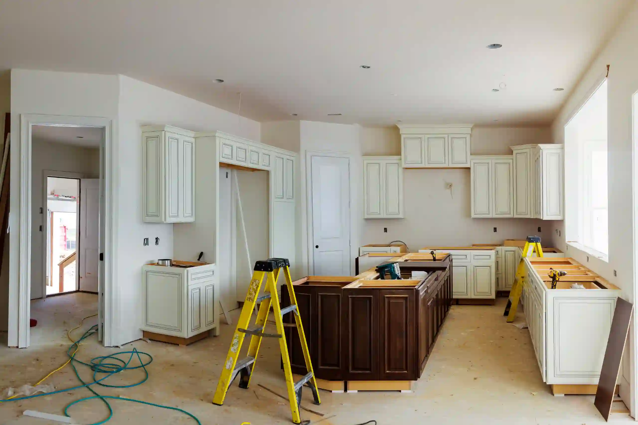 Kitchen under construction with white cabinets, a brown island, tools, a yellow ladder, and unfinished floors; sunlight comes through large windows.