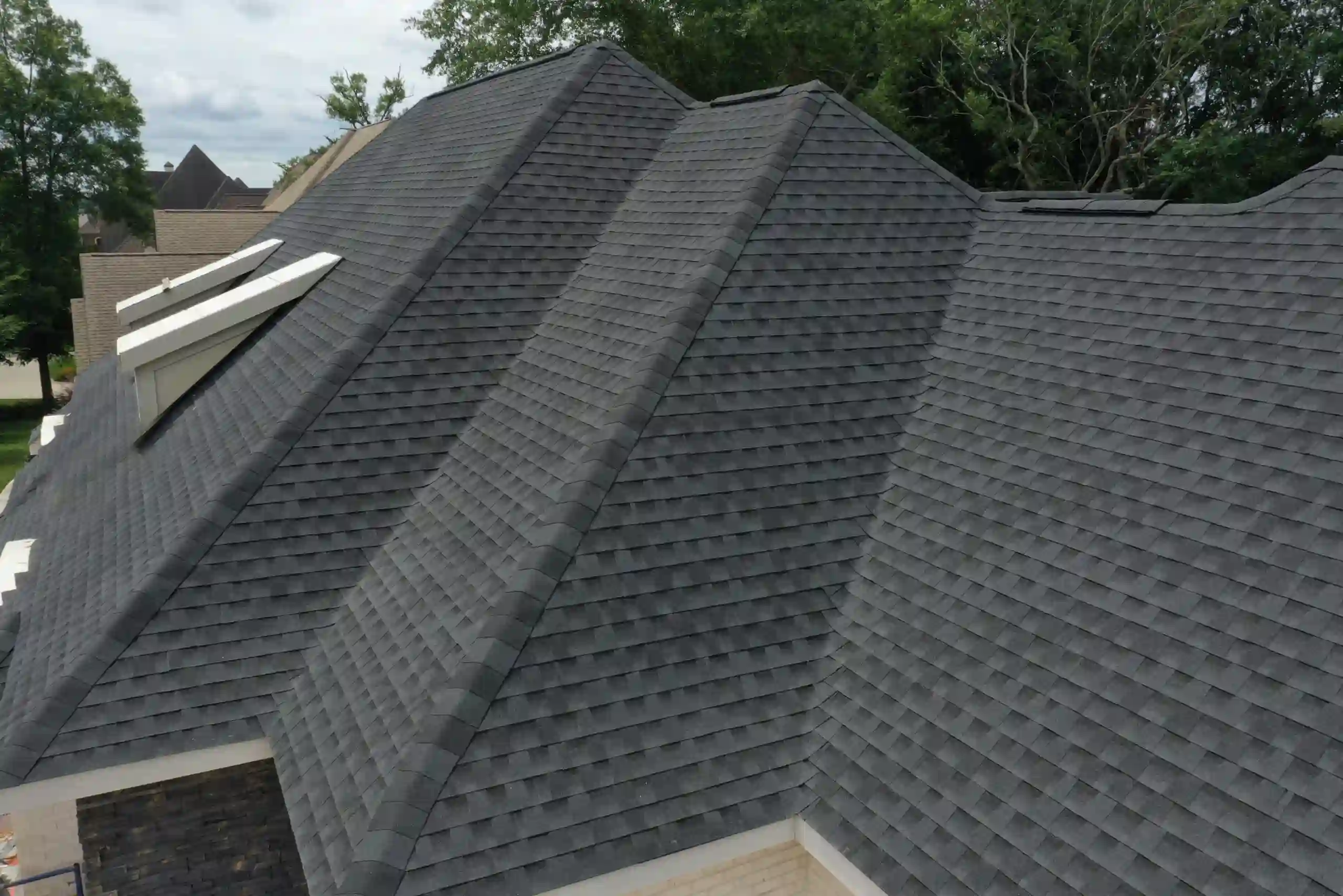 A roof with dark gray asphalt shingles and multiple ridges, viewed from above. Trees and parts of other rooftops are visible in the background.