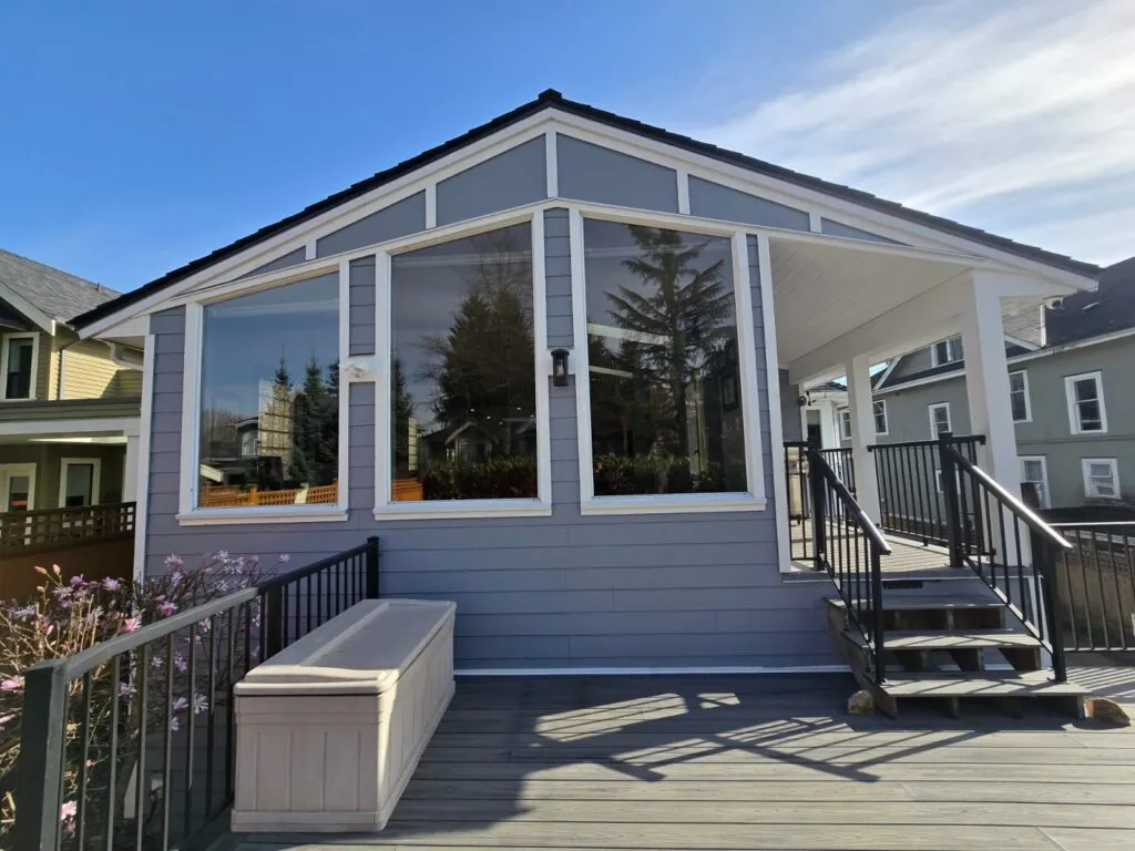 A modern porch with large windows, gray siding, black railings, steps, and a storage bench, attached to a house with trees and neighboring homes visible in the background.