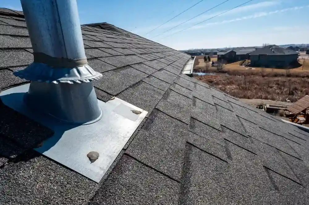 Close-up of a shingled roof with a metal vent pipe and flashing, highlighting the meticulous details often encountered during roof repair, overlooking a suburban landscape under a clear sky.