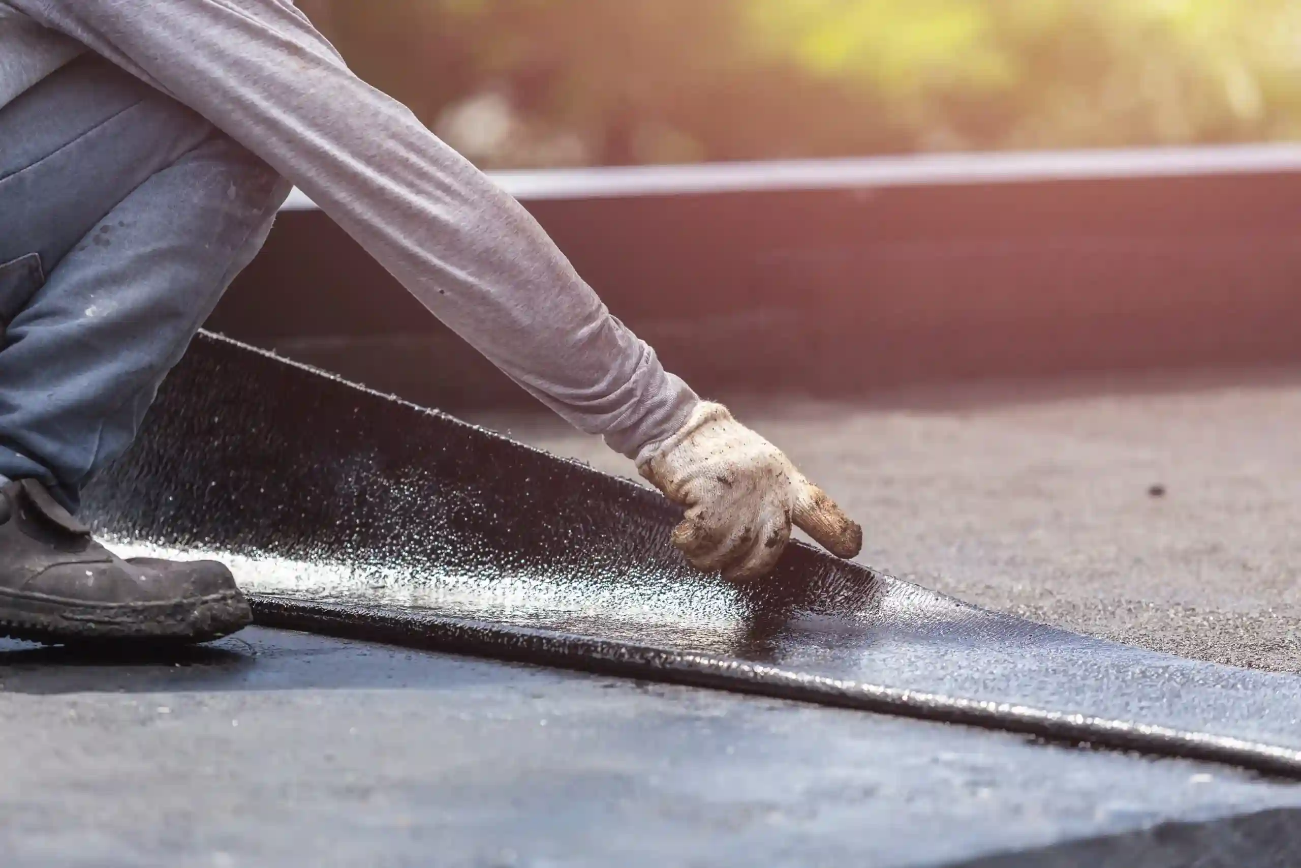 A person wearing gloves performs roof repair by installing a sheet of roofing material on a flat roof, pressing it down with their hand.