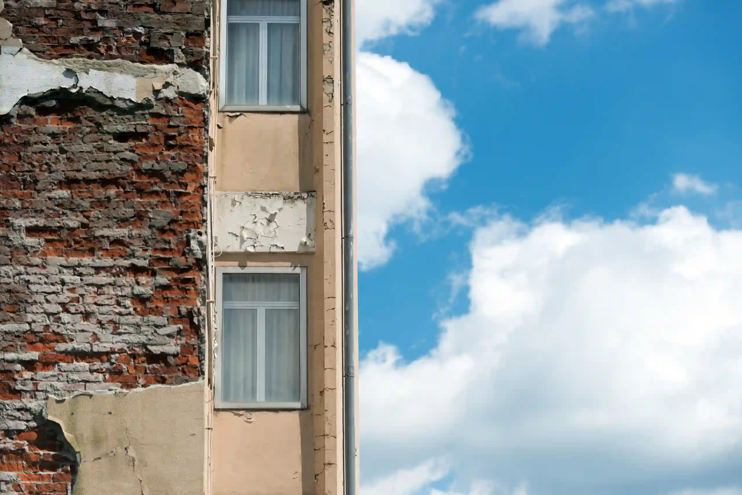 Half of a building with exposed brick and peeling stucco next to blue sky with clouds; only two windows are visible.