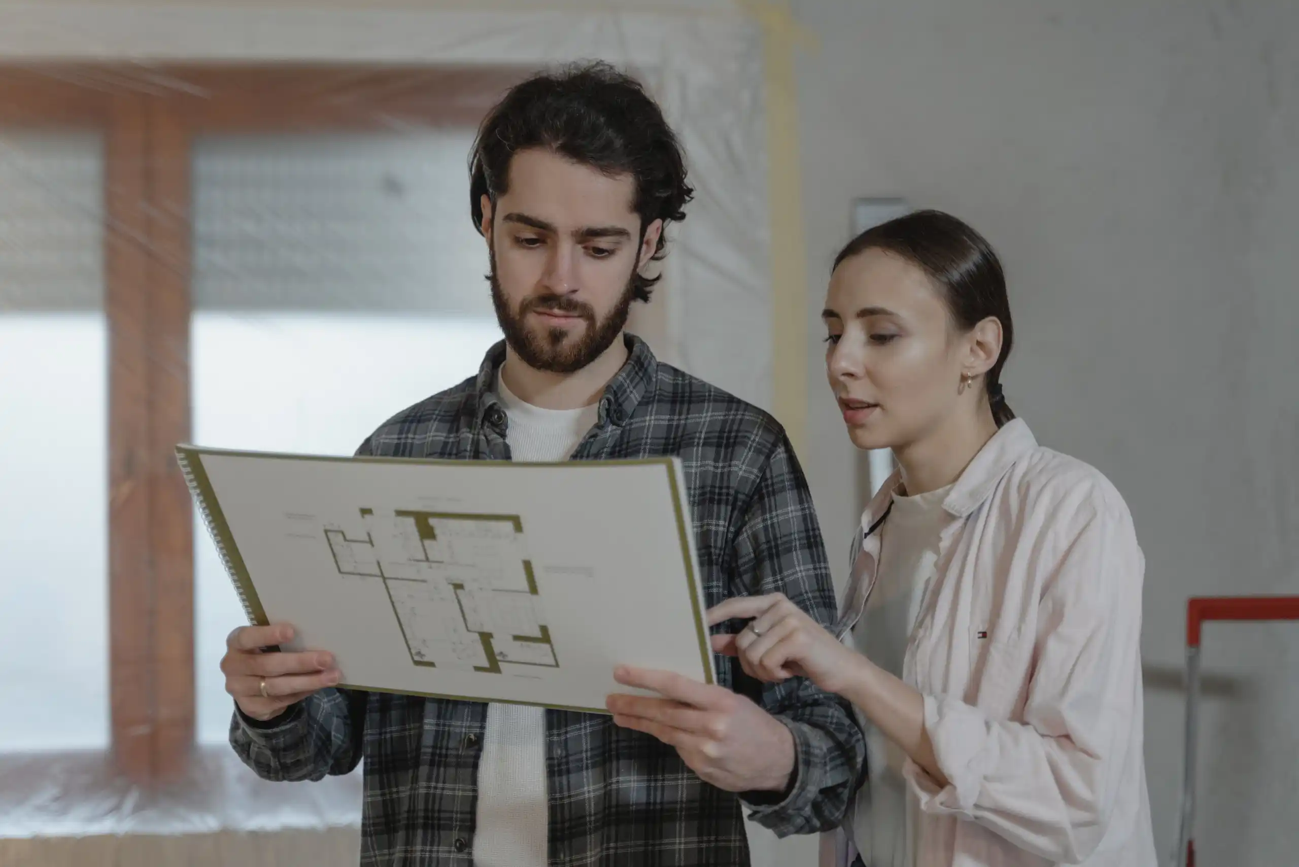 Two people stand indoors, looking at and discussing a floor plan on paper. The man holds the plan while the woman points at it. The background appears to be an unfinished room.