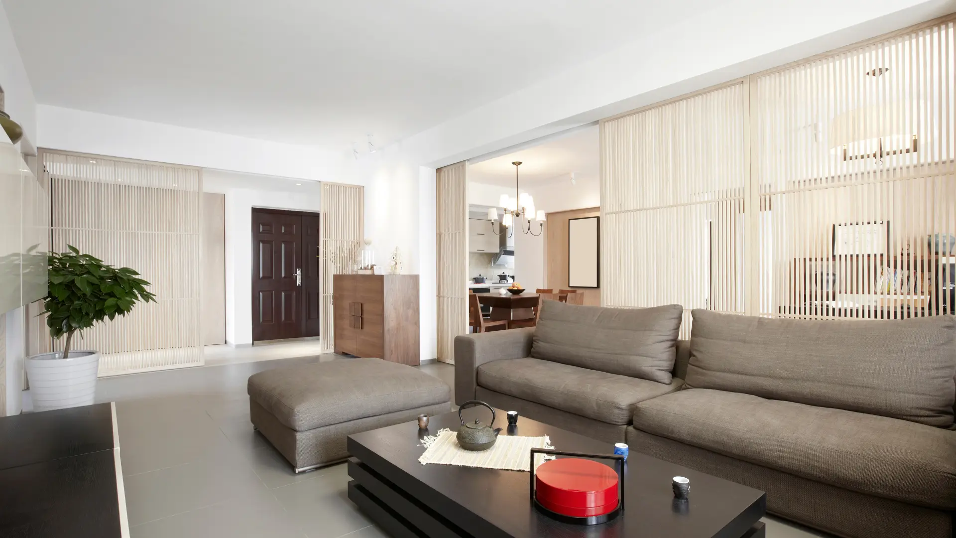 Modern living room with a gray sofa, matching ottoman, and black coffee table. Sliding wooden panels separate the dining area. A potted plant stands near a dark wooden door in the background, reflecting exquisite style seen in home renovations across Surrey.