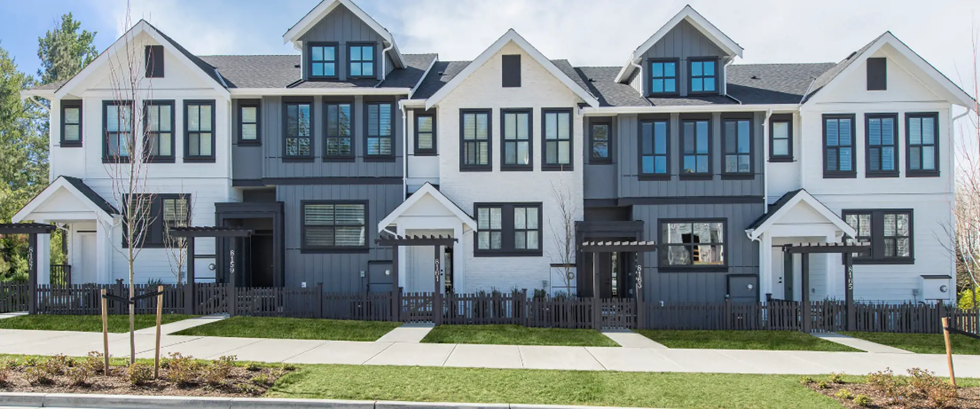 Row of modern townhouses with white and dark gray exteriors, large windows, and small fenced front yards facing a sidewalk—all featuring well-kept landscaping through regular property maintenance.