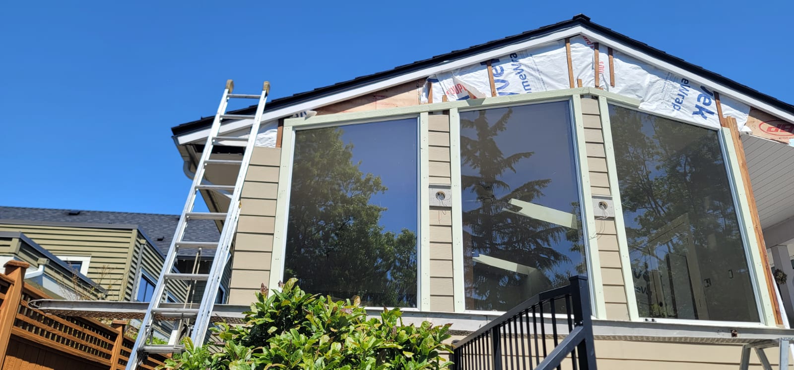 A house exterior under renovation showcases ongoing property maintenance, with a ladder leaning against the wall, exposed insulation, and large windows reflecting trees and sky.
