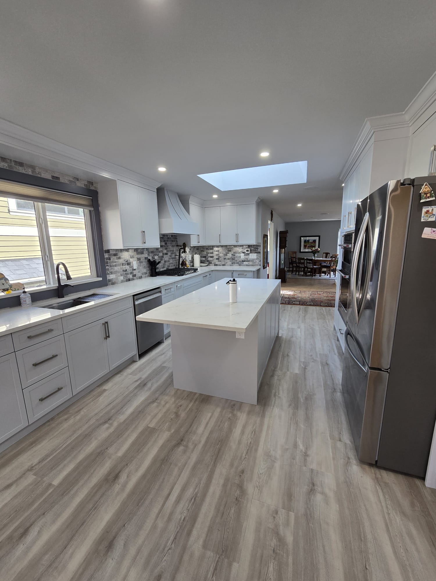 Modern kitchen with white cabinets, a large island, stainless steel appliances, gray backsplash, and wood-look flooring under a skylight—meticulously cared for with regular property maintenance.