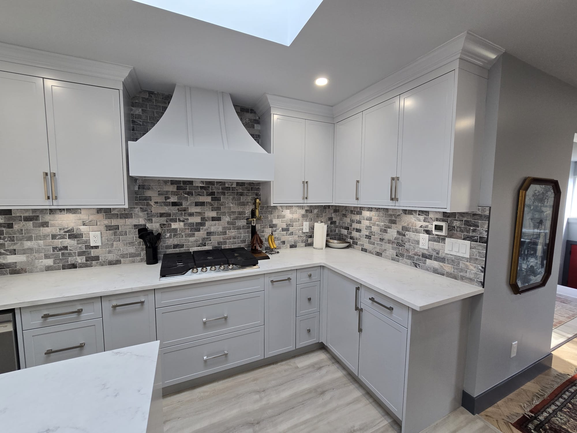 Modern kitchen with white cabinets, marble countertops, gray brick backsplash, a gas stove, and a white range hood under a skylight—showcasing excellent property maintenance throughout.
