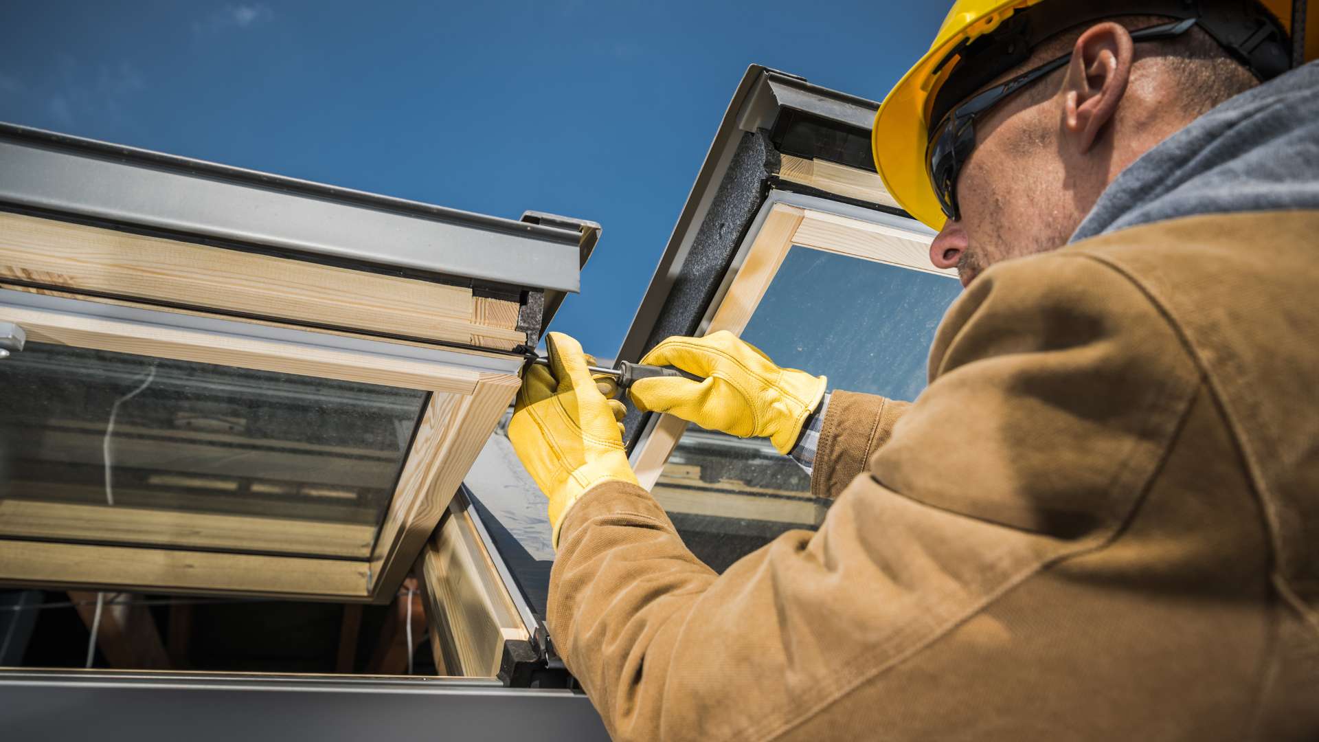 A construction worker in a hard hat and gloves installs a skylight window on a roof under a clear blue sky.