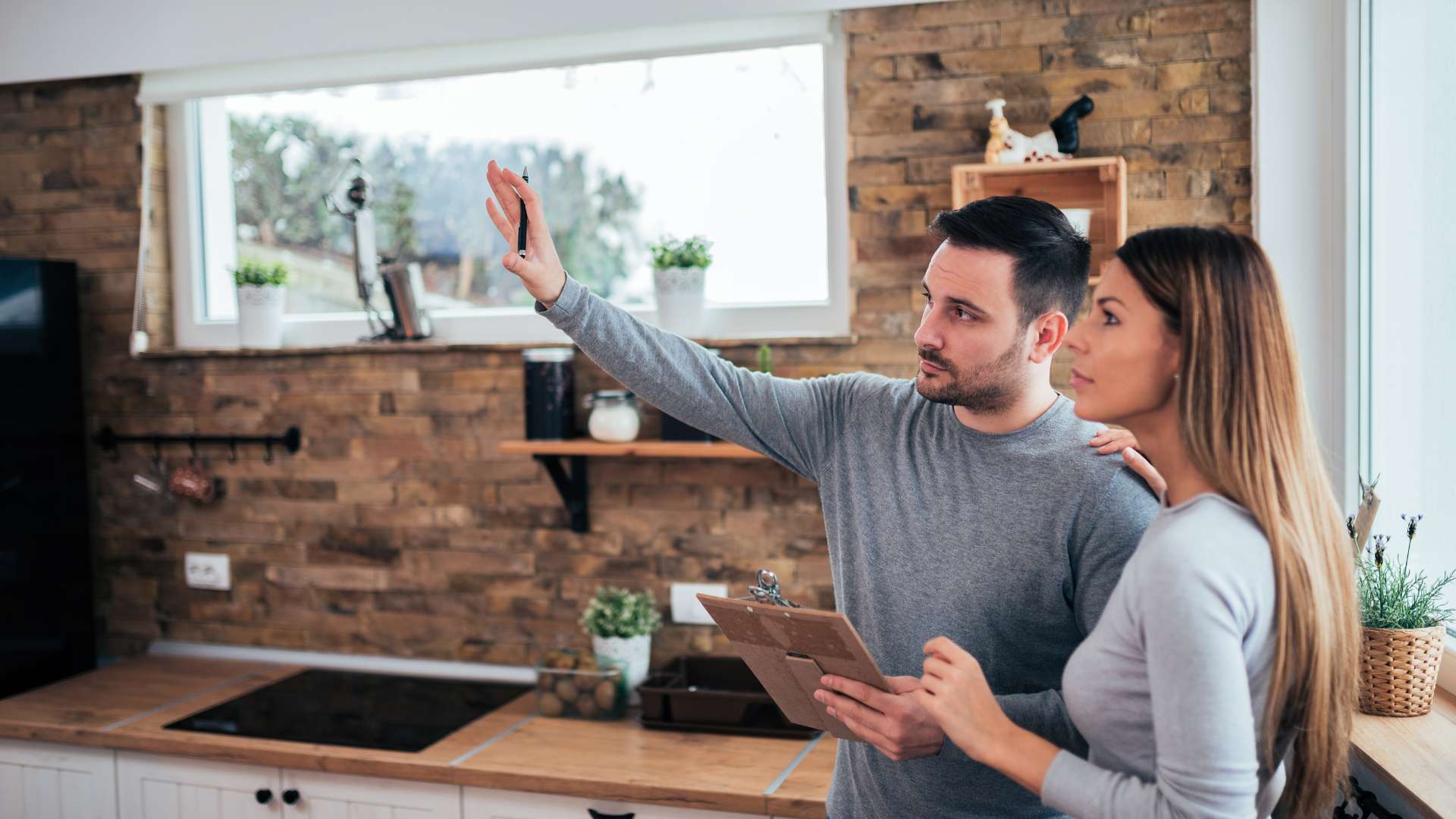 A man and woman stand in a kitchen with a brick wall; the man is gesturing upward while holding a clipboard.
