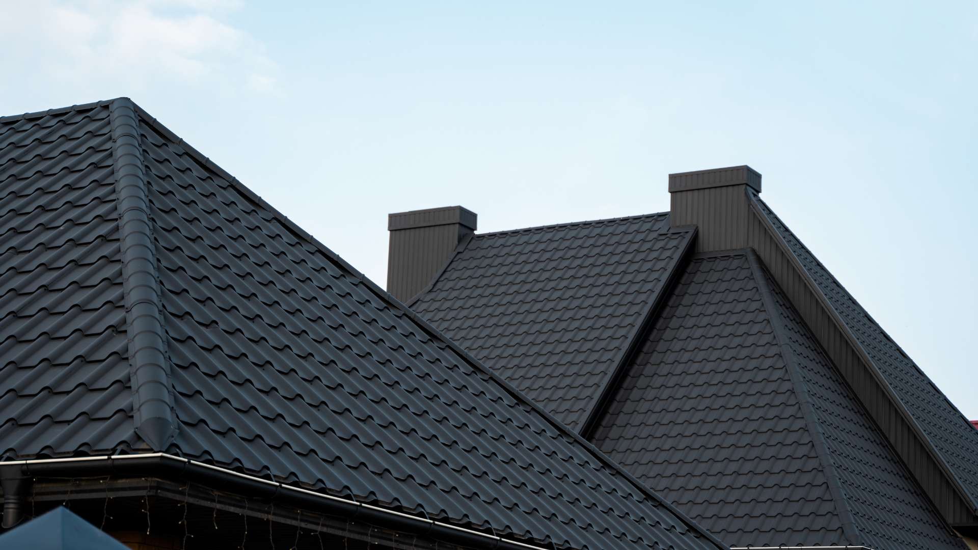 Image of a residential house with dark gray metal roofing and two prominent chimneys against a clear sky.